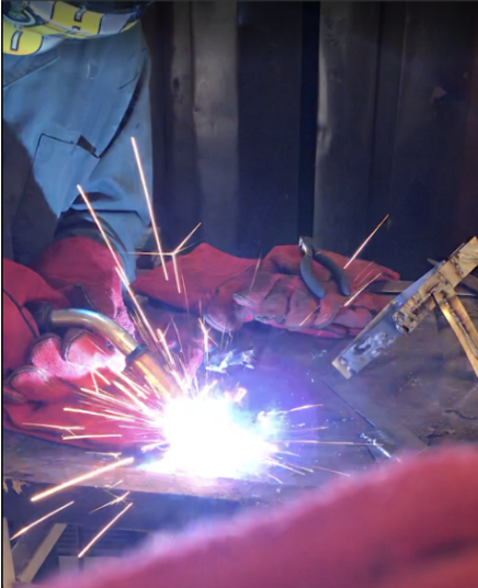 TIG welding close-up — arc and sparks during weld with gloves/clamps visible.